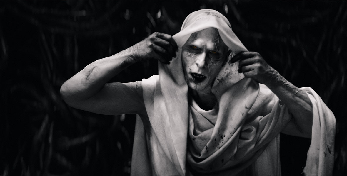 In a black-and-white production still from Marvel Studios’ Thor: Love and Thunder, actor Christian Bale, as Gorr the God Butcher, peels back his white head scarf to reveal his scarred face. His teeth are razor-sharp, and his expression is menacing.