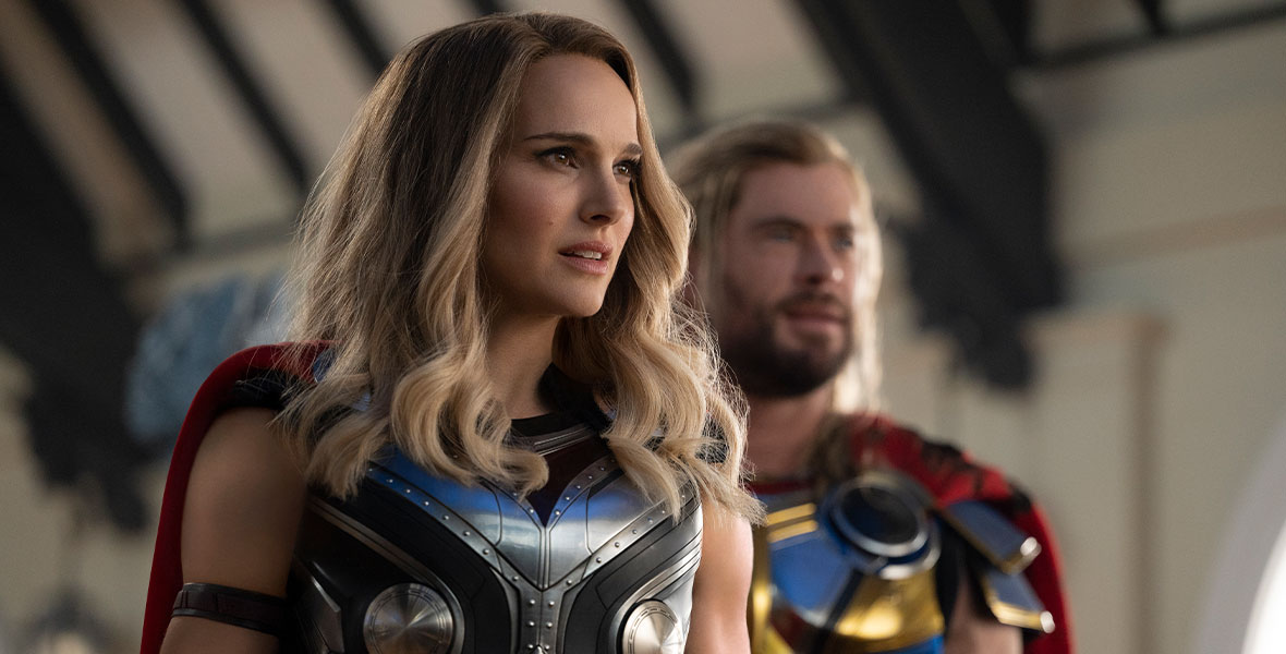In a production still from Marvel Studios’ Thor: Love and Thunder, actress Natalie Portman, as Jane Foster, wears Asgardian battle armor and a red cape. She is more muscular than ever and newly blonde. Behind her stands actor Chris Hemsworth, as Thor, who is also wearing Asgardian battle armor and a red cape.