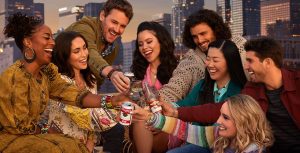 (Left to Right) In the key art for Season 4B of Freeform’s Good Trouble, actors Zuri Adele, Priscilla Quintana, Josh Pence, Cierra Ramirez, Tommy Martinez, Sherry Cola, Emma Hunton, and Bryan Craig clink drinks on the rooftop of Downtown Los Angeles’ The Coterie. They are sitting around a table at sunset, with a strand of lights hanging overhead. Above them, the Good Trouble logo appears in a pale pink font.