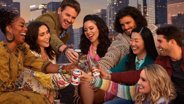 (Left to Right) In the key art for Season 4B of Freeform’s Good Trouble, actors Zuri Adele, Priscilla Quintana, Josh Pence, Cierra Ramirez, Tommy Martinez, Sherry Cola, Emma Hunton, and Bryan Craig clink drinks on the rooftop of Downtown Los Angeles’ The Coterie. They are sitting around a table at sunset, with a strand of lights hanging overhead. Above them, the Good Trouble logo appears in a pale pink font.