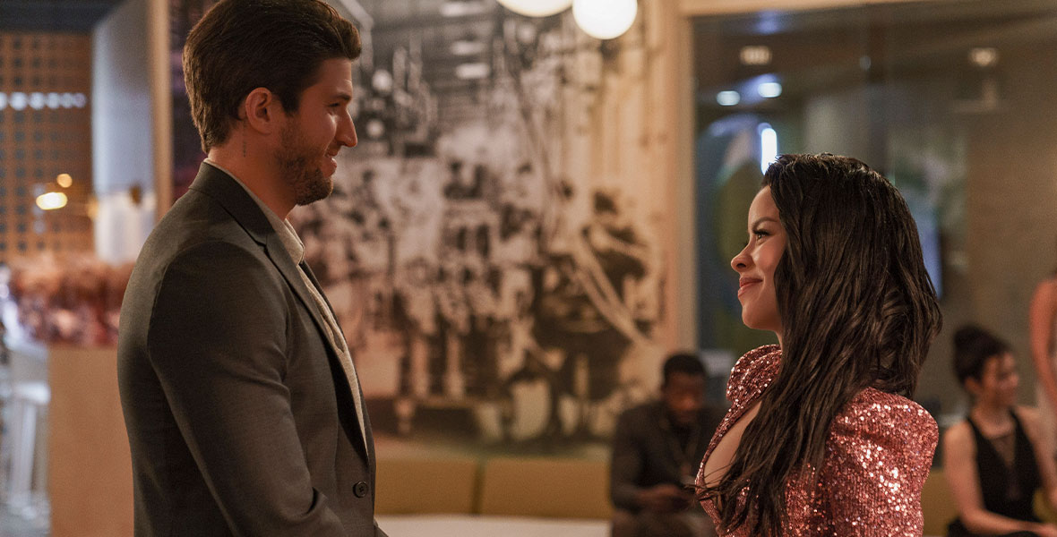In this scene from Good Trouble’s summer premiere, “What I Wouldn’t Give for Love,” Joaquin, played by Bryan Craig, stands facing Mariana, played by Cierra Ramirez, at a cocktail party. Joaquin is wearing a suit and is gripping his wrist, while Mariana is smiling back at him. She is wearing a sequin dress with a plunging neckline and holding a champagne flute.