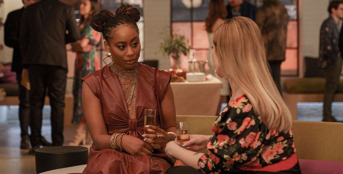 In this scene from Good Trouble’s summer premiere, “What I Wouldn’t Give for Love,” Malika (Zuri Adele) sits on a couch opposite Davia (Emma Hunton), whose back is to the camera, during a cocktail party. Malika is wearing a brown leather dress and layered necklace and bracelets, and she is holding a champagne flute. Davia is wearing a black and pink floral top and a long black skirt and is holding a champagne flute. In the background, party guests mingle in small groups.