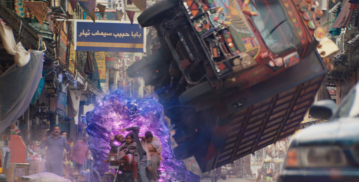 A massive chase scene is visible on the streets of Karachi, as a bus painted in colorful motifs flips on the right, Ms. Marvel’s protective illuminated shield protects a motorbike with a child, father, and mother from injury.