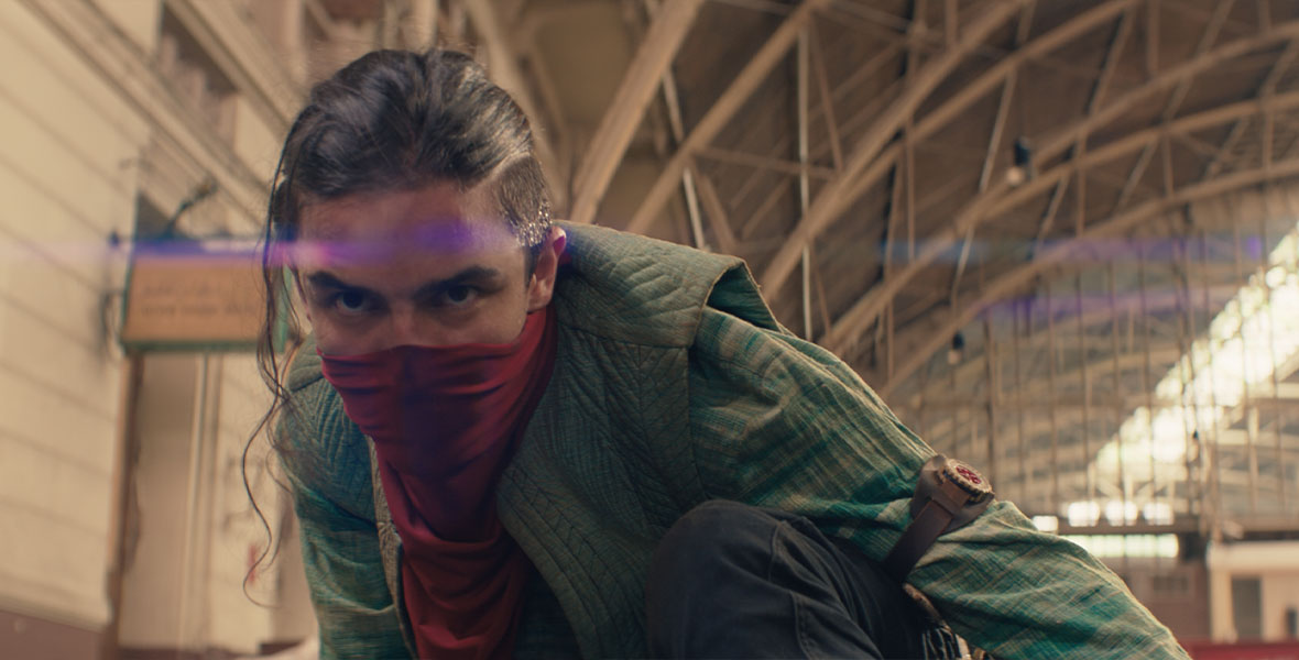 Kareem, played by actor Aramis Knight, crouches on a train station platform, with his lower face hidden behind a red sash. His stares ahead with a vigilant look in his eyes.