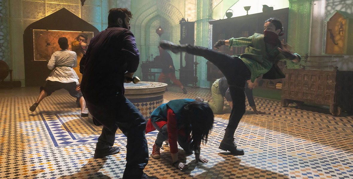 In the foreground Kareem launches a high kick at an attacker to the left, as Kamala crouches below his outstretched leg. In the background, Waleed is seen in battle against a female attacker in a group fight scene in the sanctuary of the Red Dagger society. 