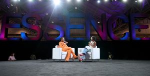 Actress Keke Palmer, in a bright orange suit, sits on a white couch and talks into a microphone while facing the panel moderator, actress and influencer Marsai Martin, who also sits on a white couch facing Palmer. Behind them both is a large display of letters in pink and blue spelling out ESSENCE.
