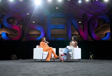 Actress Keke Palmer, in a bright orange suit, sits on a white couch and talks into a microphone while facing the panel moderator, actress and influencer Marsai Martin, who also sits on a white couch facing Palmer. Behind them both is a large display of letters in pink and blue spelling out ESSENCE.