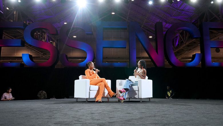 Actress Keke Palmer, in a bright orange suit, sits on a white couch and talks into a microphone while facing the panel moderator, actress and influencer Marsai Martin, who also sits on a white couch facing Palmer. Behind them both is a large display of letters in pink and blue spelling out ESSENCE.