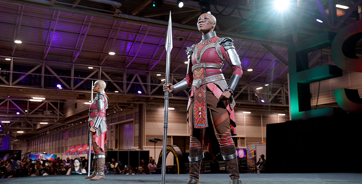 Two members of the Dora Milaje from the film Black Panther, stand proudly onstage, holding their spears in matching poses.