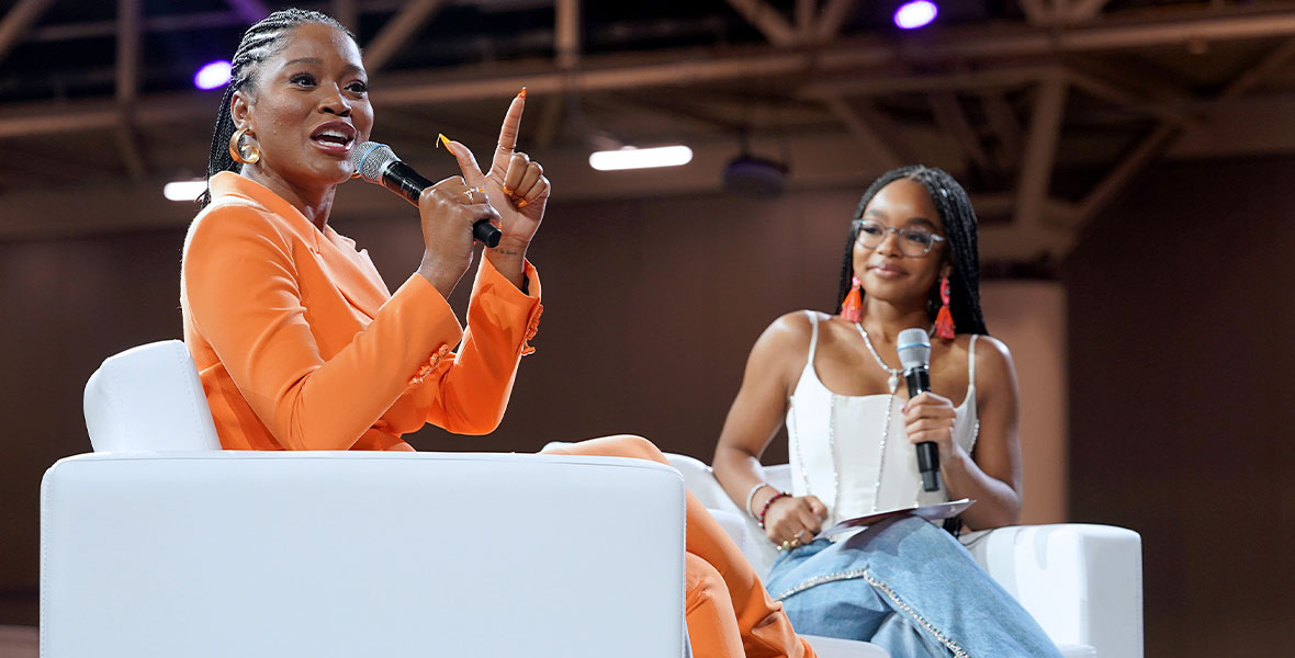 Actress Keke Palmer, in an orange suit with matching nails, speaks to the audience at a panel during ESSENCE Fest. Panel moderator, actress and influencer Marsai Martin, watches Keke speak. Both women are holding microphones and seated on white couches.