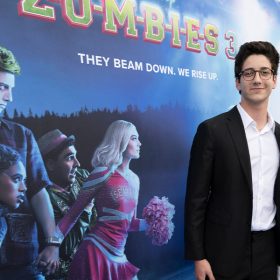 At the world premiere of ZOMBIES 3, actor Milo Manheim (left) wraps his arm around actress Meg Donnelly (right) while posing for a photo on the blue carpet. Milo wears a black suit with a white shirt and glasses, and Meg wears a white corset over a white long-sleeve gown. Meg accessorizes her look with silver earrings, necklaces, bracelets, and rings.