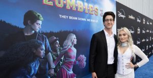 At the world premiere of ZOMBIES 3, actor Milo Manheim (left) wraps his arm around actress Meg Donnelly (right) while posing for a photo on the blue carpet. Milo wears a black suit with a white shirt and glasses, and Meg wears a white corset over a white long-sleeve gown. Meg accessorizes her look with silver earrings, necklaces, bracelets, and rings.