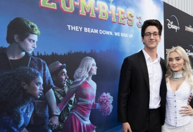 At the world premiere of ZOMBIES 3, actor Milo Manheim (left) wraps his arm around actress Meg Donnelly (right) while posing for a photo on the blue carpet. Milo wears a black suit with a white shirt and glasses, and Meg wears a white corset over a white long-sleeve gown. Meg accessorizes her look with silver earrings, necklaces, bracelets, and rings.