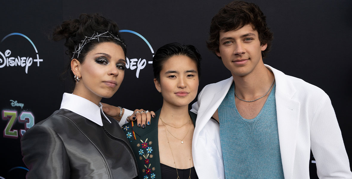 At the world premiere of ZOMBIES 3, co-stars Kyra Tantao (left), Terry Hu (center), and Matt Cornett (right) wrap their arms around each other for a photo against the movie’s branded backdrop, which features the logos for ZOMBIES 3 and Disney+. Kyra is wearing a futuristic jacket with a white collar, and she has dozens of pins in her hair; Terry is wearing a floral embroidered blazer and a black bandeau top, along with delicate gold jewelry; and Matt is wearing a white blazer and over an icy blue tank top.