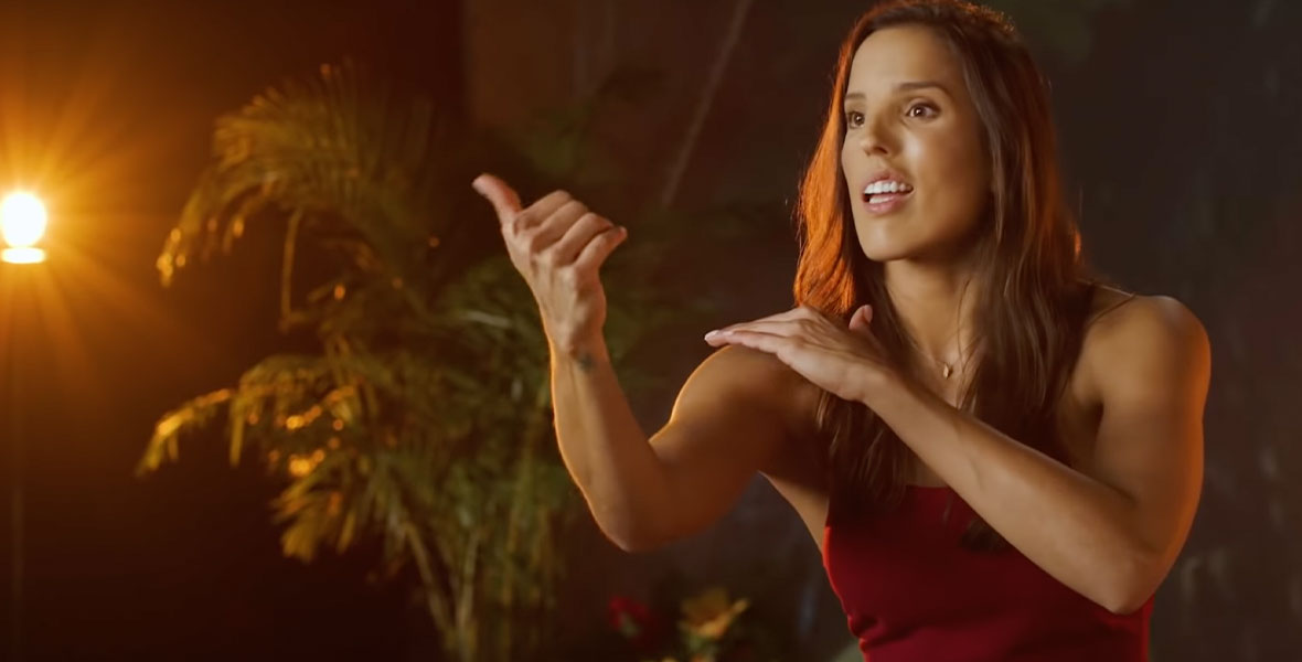 : Deaf artist Sarah Tubert performs “How Far I’ll Go” from Walt Disney Animation Studio’s Moana in ASL. Tubert has long brown hair and is wearing a deep red tank top. She is standing in front of a large screen that is playing the “How Far I’ll Go” sequence from Moana in time with her signing. There are large palm trees and flowers surrounding her on set and she is looking off into the distance as she performs.