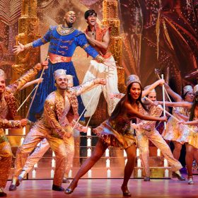 Michael James Scott performs as Genie with his arms outstretched next to Michael Maliakel performing as Aladdin against a gold-covered set. In front of them, a row of performers in gold dance with canes.