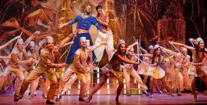 Michael James Scott performs as Genie with his arms outstretched next to Michael Maliakel performing as Aladdin against a gold-covered set. In front of them, a row of performers in gold dance with canes.