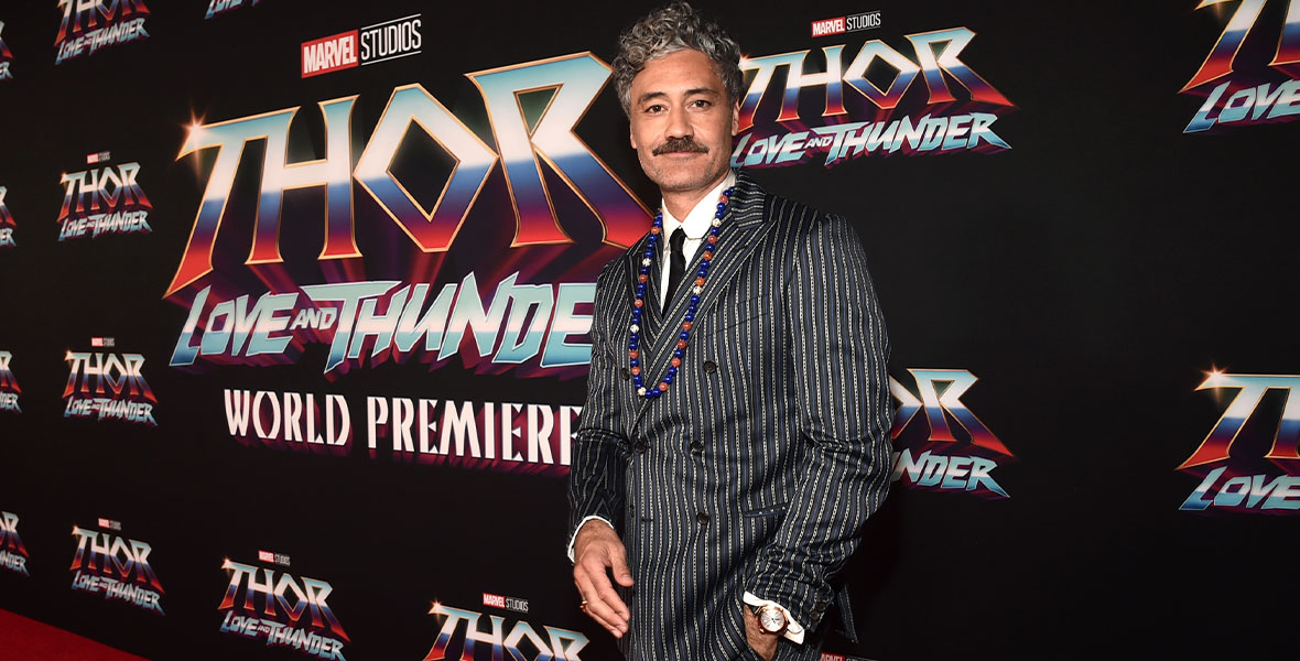 Taika Waititi stands on the red carpet for Thor: Love and Thunder and poses in front of a step-and-repeat for the film. He has curly salt and pepper hair, a mustache, and is wearing a black and white pinstripe suit. Around his neck is a large necklace made up of red, blue, and white spherical beads. He has one hand casually hanging in his pocket and he is smiling with no teeth at the camera.