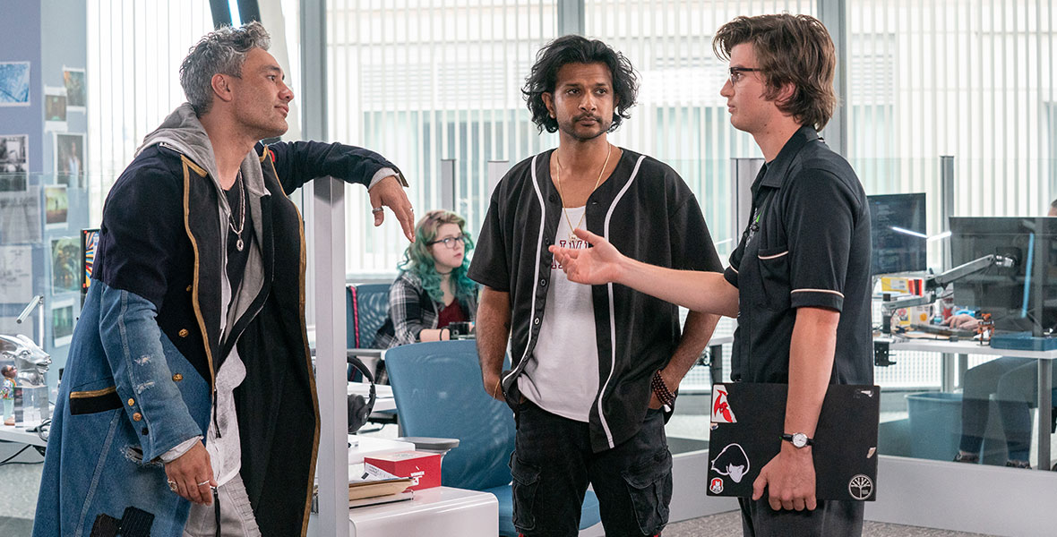 Taika Waititi, Utkarsh Ambudkar, and Joe Keery stand facing each other in a sleek, modern office setting, in Free Guy. Waititi has on a long trench coat made of multiple materials, including denim. He is resting casually against the side of a cubicle and is looking at Keery, whose character is speaking. Keery is holding a laptop at his side and is wearing a black polo shirt. In the middle is Ambudkar who is wearing an unbuttoned baseball jersey over a white tank top and black jeans with red lining.