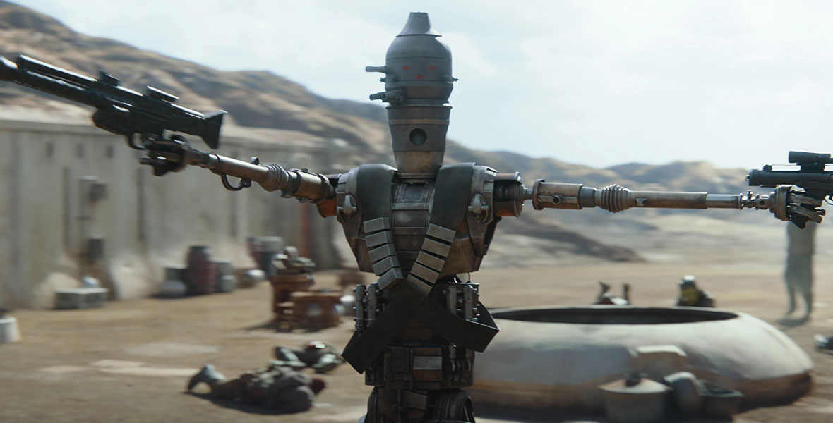 The IG-11 droid from The Mandalorian stands front and center with both mechanical arms outstretched holding blasters. Behind the droid there are bodies on the ground and one stormtrooper approaching. IG-11 wears a double-breasted holster across the center of its body.