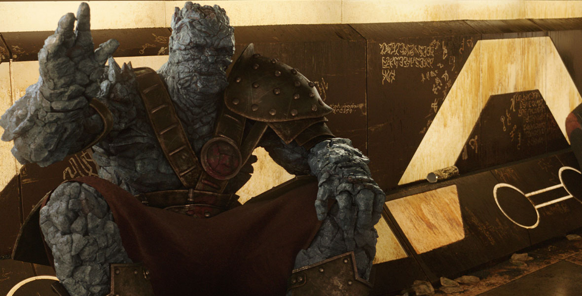 Korg (Taika Waititi), a gigantic greyish blue rock creature called a Kronan, sits on the ground against a wall and waves one hand pleasantly towards the camera in Thor: Ragnarok. He is wearing a kilt and large metal armor across his body and over his left shoulder. He is leaning against a brown wall with white accents and details and graffiti written in an alien language.