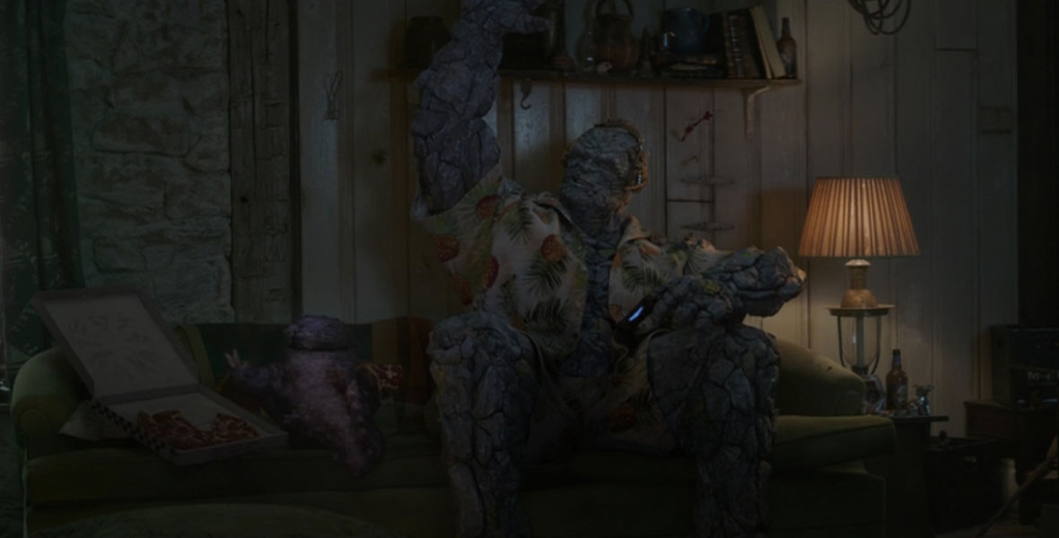 Korg (Taika Waititi) sits on Thor’s (Chris Hemsworth) couch in a Hawaiian shirt, holding a video game controller and wearing a headset, in Avengers: Endgame. He is raising his right arm in a wave towards characters off-camera who are greeting him. To his left is another alien creature, Miek, a worm-like being from Sakaar. To Miek’s left is an open box of pepperoni pizza.