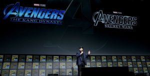 Kevin Feige on stage presenting the Marvel live action presentation. He is in a blue suit jacket with black shirt undernearth and black pants. He also has a black baseball hat on and blue and white sneakers. Behind him on a huge screen are the logo title images for Avengers: The Kang Dynasty and Avengers: Secret Wars.