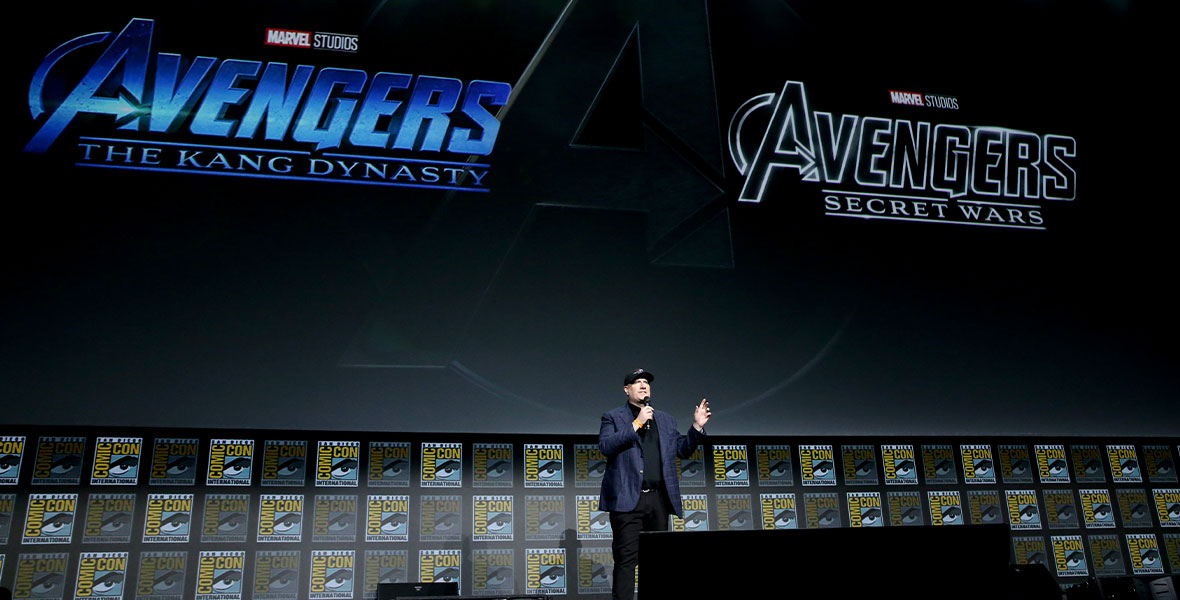 Kevin Feige on stage presenting the Marvel live action presentation. He is in a blue suit jacket with black shirt undernearth and black pants. He also has a black baseball hat on and blue and white sneakers. Behind him on a huge screen are the logo title images for Avengers: The Kang Dynasty and Avengers: Secret Wars.