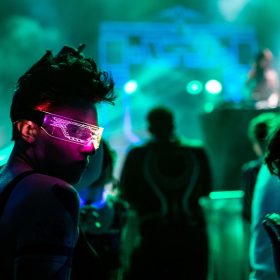 Tron fan with pink light-up glasses looking over their shoulder. The photo is dark with silhouetted figures of other fans and DJ Qrion in the back with green and blue foggy lights.