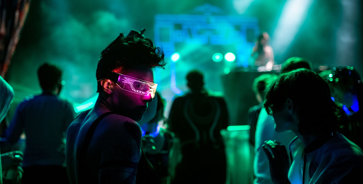 Tron fan with pink light-up glasses looking over their shoulder. The photo is dark with silhouetted figures of other fans and DJ Qrion in the back with green and blue foggy lights.