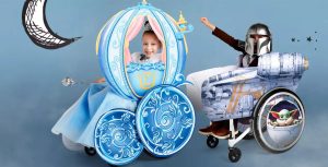 A child dressed as Cinderella sits in a wheelchair decorated like Cinderella's coach. To the right of her, a child dressed as The Mandalorian raises his fist in the air as he sits in a wheelchair decorated to look like The Mandalorian's ship, the Razor Crest. A depiction of Grogu sitting in his hovering pram covers the chair's wheel.