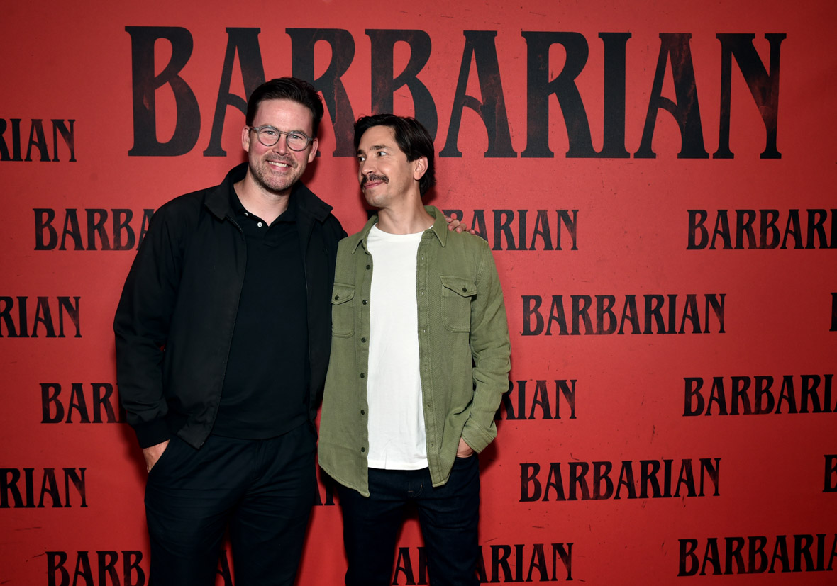 Zach Creggar and Justin Long standing in front of a red step-and-repeat with the Barbarian logo all over it. Creggar is in all black (jacket, shirt, pants) and is wearing round glasses. Justin Long is smiling at Creggar. He has shaggy black hair and a mustache. He is wearing a green denim jacket over a white tee shirt and black pants.