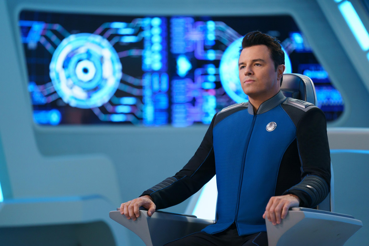 Seth MacFarlance in The Orville. He is on a spaceship in a captain’s chair wearing a blue and black space uniform with silver details on the shoulders and chest lapel. He is looking off camera with a stern look on his face and behind him are space-like graphics on a wall or screen.