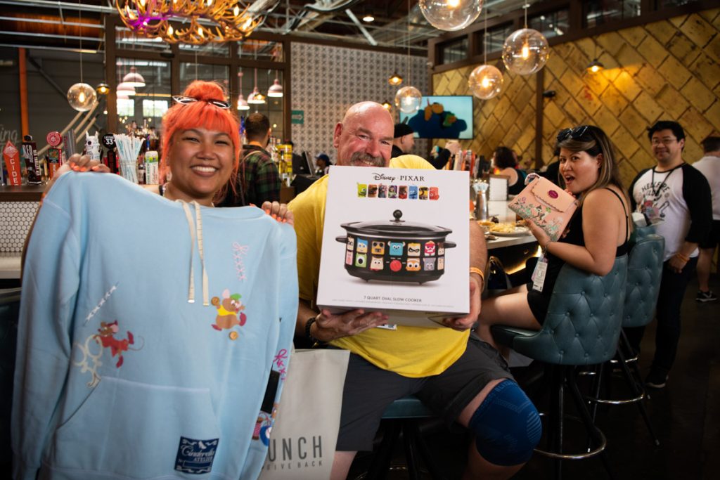 A fan smiling with bright orange hair and sunglasses is posing with her light blue Cinderella sweatshirt featuring Gus and Jaq. Sewing-inspired details such as stitches and measuring tape draw strings that she won at the BoxLunch prize wheel. The fan to the right of her is holding a boxed crockpot that is black with squared Pixar characters covering the crockpot. The fan is wearing a bright yellow shirt and grey shorts with a blue knee brace. The fan behind them is gasping in awe at the pink Bambi purse that she won at the wheel too. She is dressed all in black. They are all standing and sitting by the Punchbowl Social circular bar featuring bubble type lamps and green chairs.