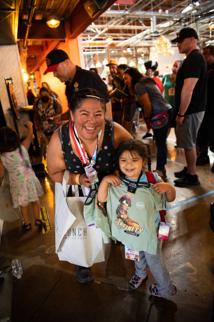 A fan smiling posing with her little one after winning prizes at the prize wheel. The fan is wearing a sparkly headband, a black tank top with Marvel Super Hero circle logos. They are holding a mini–Soul Pop Funko figure as well as a canvas bag with BoxLunch’s logo. Next to them is a younger fan with ponytail braids, wearing a light blue shirt, grey sweatpants and multicolor shoes. They are smiling holding up their light green kid’s shirt featuring the “Infinity Cone” company with the Infinity gauntlet holding a vanilla ice cream cone topped with chocolate syrup and gum drops resembling the infinity stones.