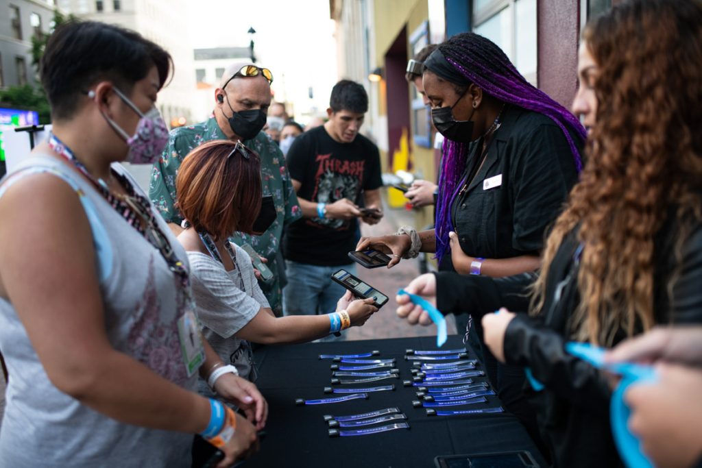 Guests standing in line to check in for the event outside the House of Blues. Four guests on the left of the table are patiently waiting. The fan closest to the camera is wearing a white tank top and has a white face mask with pink patterned design. The next fan is showing their ticket to the staff. They have short brown hair with sunglasses on their head, a black face mask and white T-shirt. The fan behind has a black face mask and green button-up shirt with sunglasses on their head. The fan behind is getting their ticket scanned by staff. The fan has a black T-shirt with a central design and blue jeans. The table is covered with a black tablecloth with blue glow wristbands. On the right side of the table are two staff members wearing black House of Blues shirts and passing out blue drink tickets.