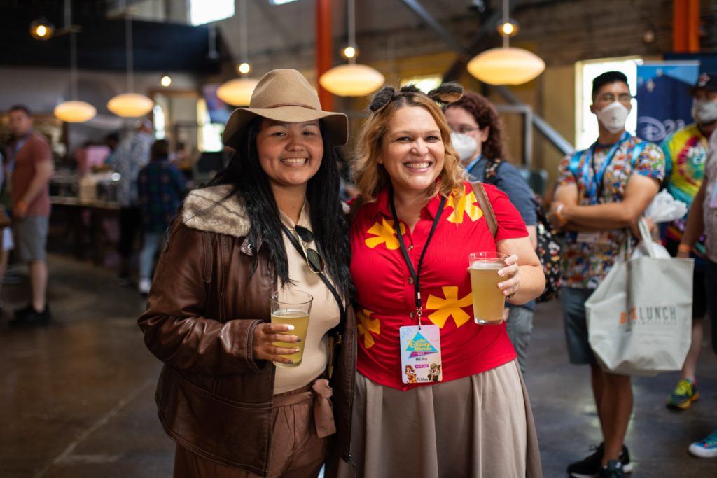 Two fans smiling with their beers dressed as Chip and Dale Rescue Rangers. The fan on the left has dark hair, brown fedora, brown leather jacket, brown shorts and tan shirt. Sunglasses hang across their neck with a black satchel. The fan on the right has mid-length reddish hair, chipmunk ears, red Hawaiian shirt with yellow flowers with a light brown skirt and brown backpack. There are multiple fans mingling around in the background.