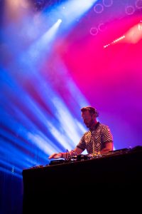 DJ Jason Bently, former music director of KCRW and music supervisor for the Tron: Legacy soundtrack stands behind black DJ table and turn tables on top. DJ Bently is standing on a stage with pink and purple back lighting and blue streak lights on the left of the photo. DJ Bently is wearing over-the-head headphones and a checkered black and white polo.