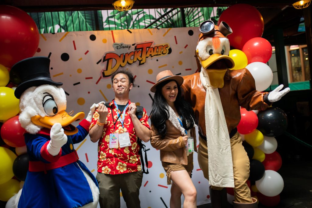 Two fans posing with Scrooge McDuck and Launchpad McQuack. Scrooge is on the far left with his hand on the fan to his right. Scrooge has white feathers, a yellow beak, black top hat, glasses sitting on his beak in front of his eyes, and a royal blue coat with red belt and cuffs and yellow buttons. The fan posing with Scrooge is making their best duck face with arms bent up. This fan is wearing a red Hawaiian shirt with yellow flowers and dark green pants with a black backpack. The fan next is wearing a brown fedora, brown leather jacket, white shirt and brown shorts. This fan is smiling wide and has long dark hair side-hugging Launchpad. Launchpad has his arm around the fan and other arm is presenting out. Launchpad is tall and muscular with white feathers, orange beak, and tuft of orange hair hanging out of his brown leather pilot’s hat. On top of his hat are his aviator glasses. Launchpad has a brown leather jacket, tan pants, and an off-white scarf with knee high brown boots. The photo backdrop that is white with red, yellow and black confetti shapes, there are red, white, yellow, and black balloon towers on either side and a DuckTales logo in the top center of the backdrop.