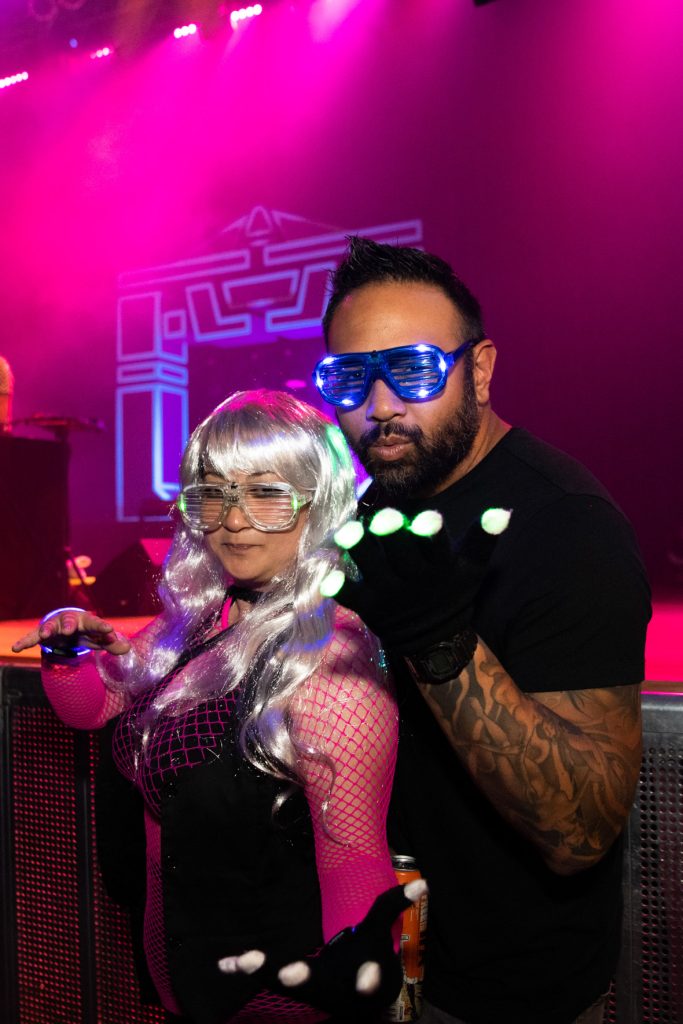 Two fans posing in game fight poses in front of the DJ stage. The fan on the left has long silver hair with silver slated glasses. The fan is wearing a neon fishnet long sleeve body suit over a black bralette which is covered with a black vest. The fan on the right has blue light up slated glasses and a black T-shirt with a tattoo-sleeved arm. Each fan has a black glove with light-up white fingertips. In the background, the Recognizer prop in black and blue is featured under pink lights.