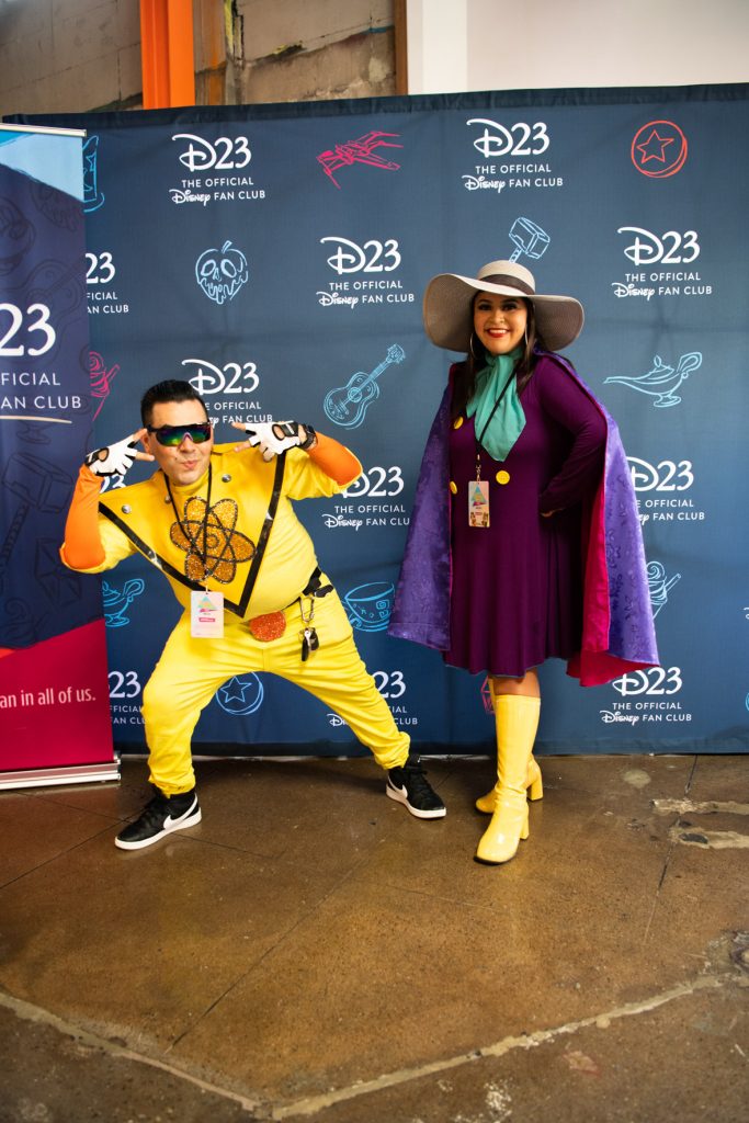 Two fans posing in front of the D23 step and repeat photo backdrop. This backdrop is navy blue with the white D23 logo intertwined with light blue and red sketches of Disney artifacts including the Pixar ball, poison apple, Coco’s guitar, teacup, Dole Whip, genie lamp, Thor’s hammer, and Star Wars ship. On the left of the backdrop is a vertical D23 banner with light blue paint stripe at the top, navy blue background with navy blue sketches that were on the photo backdrop and white D23 logo in the middle. At the bottom is a dark pink paint stripe and reads “the fan in all of us”. The fan on the left is dressed in their best Powerline costume. They are wearing a bright yellow body suit with gold nucleus in the center. Under the body suit is orange long sleeved shirt in addition to white gloves and black tennis shoes. They are posing with two peace signs pointed towards their face. The fan to the right of them is dressed in their best Darkwing Duck costume. They have a large grey floppy hat with black ribbon across the middle. The fan has long dark hair, hoop earrings and teal scarf. They are wearing a bright purple long sleeve dress with gold buttons, a purple cape lined magenta on the inside and high-top yellow leather boots. They are smiling with their hands on their hips.