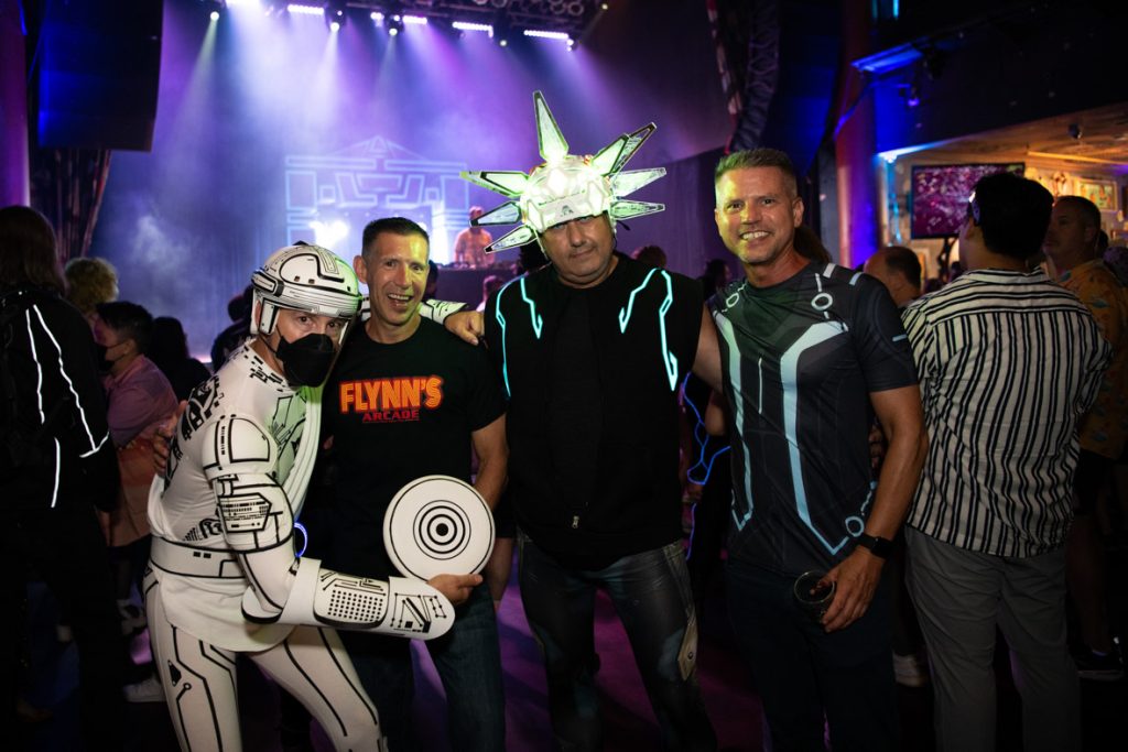Four fans standing in a line on the dance floor. On the left, the fan is wearing a black face mask with a complete Tron player inspired outfit. The white body suit is outlined with back lines showcasing a video game inspired look. The fan has a helmet that matches the suit as well as armor on his right arm and identity disk which the fan is holding, all in the same pattern as the body suit. The fan next to them is smiling and wearing a black T-shirt featuring the Flynn’s Arcade logo in orange and red with blue jeans. The next fan has a white light-up helmet with nine spikes on connected. The fan is wearing a black T-shirt with light-up lines and blue jeans. The fan on the right is smiling and wearing a black T-shirt with blue and grey designs resembling a Tron Legacy player and blue jeans. In the background are multiple fans dancing on the dance floor, and DJ Jason Bentley playing on stage. Behind DJ Bentley is the Tron Recognizer prop in blue and black. The lights on stage are white, purple and blue.