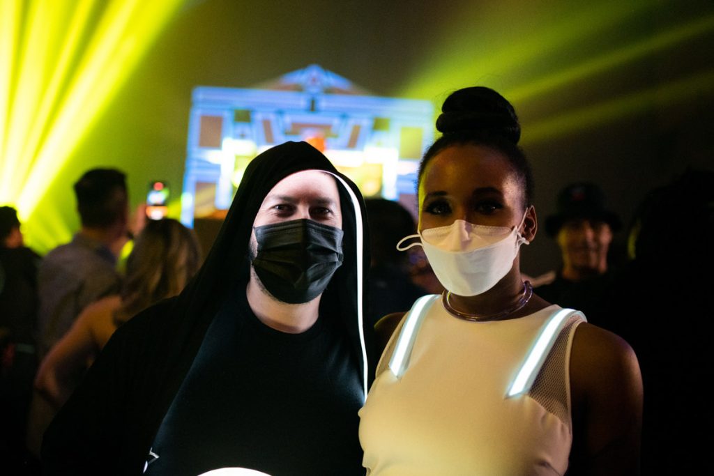 A close-up photo of two fans. The fan on the left has on a black hoodie with light-up interior, black face mask, and black T-shirt. The fan on the right has their dark hair pulled in a bun, white face mask, hard plastic choker and white tank top dress with light-up stripes on the shoulders. In the background are fans dancing and the Tron Recognizer prop in blue and black blurred. The lights in the background are yellow and green.
