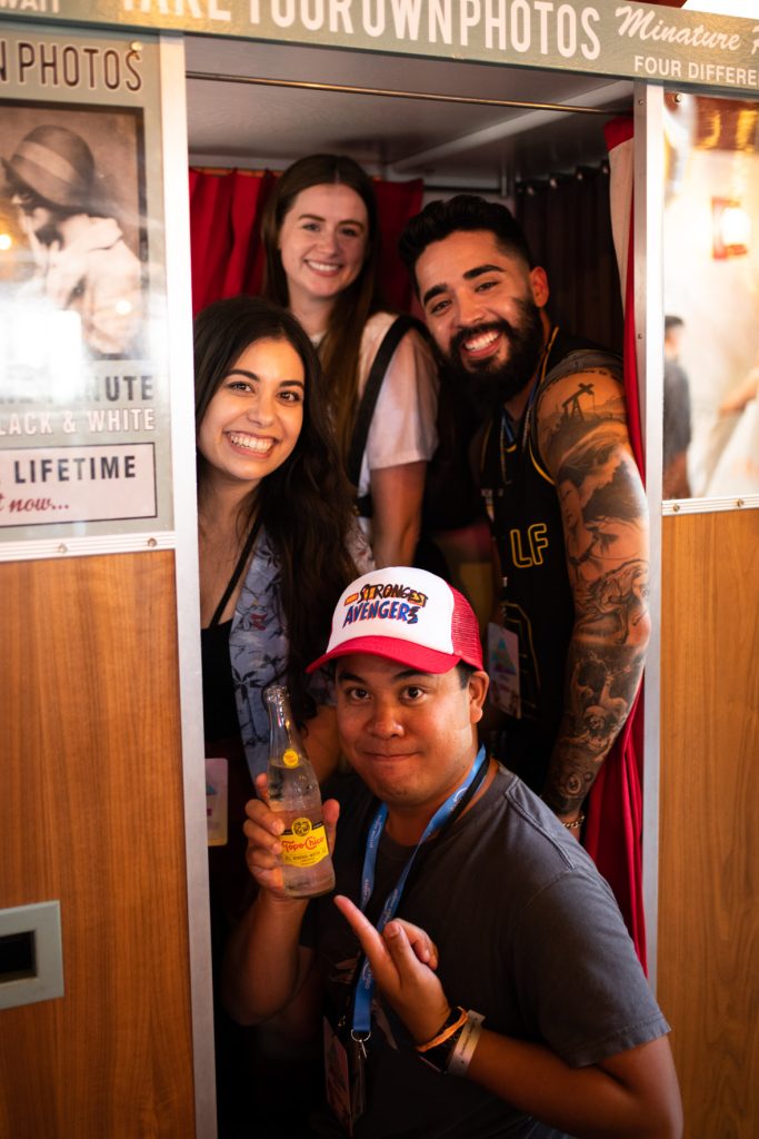 Four fans in the doorway of a retro photobooth. The photobooth is wood-paneled with black and white photos at the top of each door. The fans are posed inside the doorway of the photobooth with one standing in the back, two to the sides crouching and one in the front kneeling. The fan in the back has long brown hair and is wearing a white shirt. The fan crouching on the left has a blue Hawaiian shirt, long dark hair, and black tank top. The fan crouching to the right has a tattoo sleeved arm and is wearing a basketball jersey. The fan kneeling in the front has a snapback hat that is white and red. It reads Strongest Avengers. They are also wearing a grey T-shirt. The fan is pointing to a clear Topo Chico with yellow logo and red lettering.