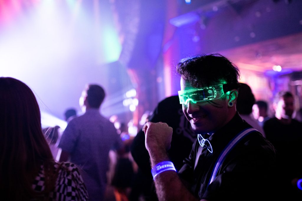Close-up on fan smiling holding up their arm showing off their blue light up wristband with the D23 Derezzed logo, green light-up glasses, and light-up bowtie and suspenders. The fan has black solid earrings and black button-up shirt. In the background are fans standing on the dance floor with purple, white and blue lights blurred.