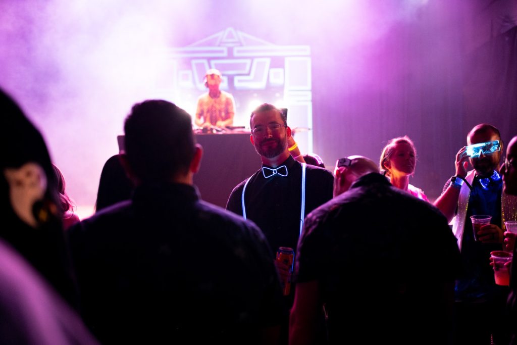 Photo focused on fan smiling with glasses, light-up bowtie and suspenders and long-sleeve black button-up shirt. The fan is surrounded by fans mingling on the dance floor. Behind the fan is DJ Jason Bently spinning on the black DJ table with the Tron Recognizer prop behind him in black and blue. The lights are foggy and in purple and white.