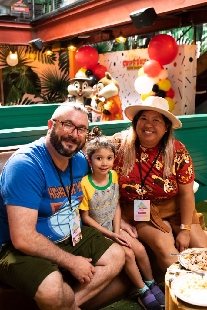 A family of three sitting on a brown bench against a teal wood background. The fan on the left has glasses, a blue T-shirt and dark green shorts. This fan’s arm is around the youngest fan in the middle. This fan’s brown hair is pulled back into a bun, has a grey DuckTales shirt with green color and yellow sleeves, and purple tennis shoes with blue socks. The fan on the right has long brown hair, a tan fedora hat, red shirt featuring Chip and Dale Rescue Rangers’ characters and brown shorts. In front of them are two plates of half-eaten food. Behind the fans is the photobooth location, Chip and Dale are meeting with a fan in front of the photobooth location. The walls behind the chipmunks are white with large green palm leaves. Above the walls are a large rust colored beam with black speakers hanging off them. Above that is the gate that protects the second floor.