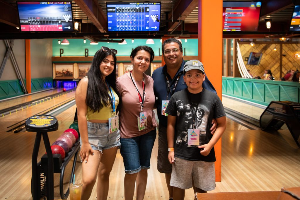 A family of four smiling in a line against a background of the bowling alley. The fan on the left has long black hair with sunglasses on their head. They are wearing a yellow cropped tank top with light denim shorts. The fan next in line has black hair pulled back, a light red shirt and denim mid-length shorts. The fan next in line is wearing glasses, a dark navy polo shirt and grey cargo style shorts, their arm is around the fan next in line. This fan has a grey baseball hat with the Comic Con logo. They are wearing a black shirt featuring Venom in black and white. They have light grey shorts on. All fans are wearing pink and blue ombre credentials featuring Chip and Dale Rescue Rangers and block shapes in 90’s style. The bowling alley has light wood floors, with black light on the pins. It is lined with teal wood walls and has a large orange beam on the right side. There are four lanes and above each lane is a tv with the teams scores on a blue or red background.