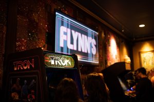 Flynn’s arcade logo projected on a large TV in white rounded block letters outlined in red. Background is light and dark blue strips with a thin red lined boarder. Below the TV Is a retro cabinet A Tron video game is visible with fans silhouetted playing it.