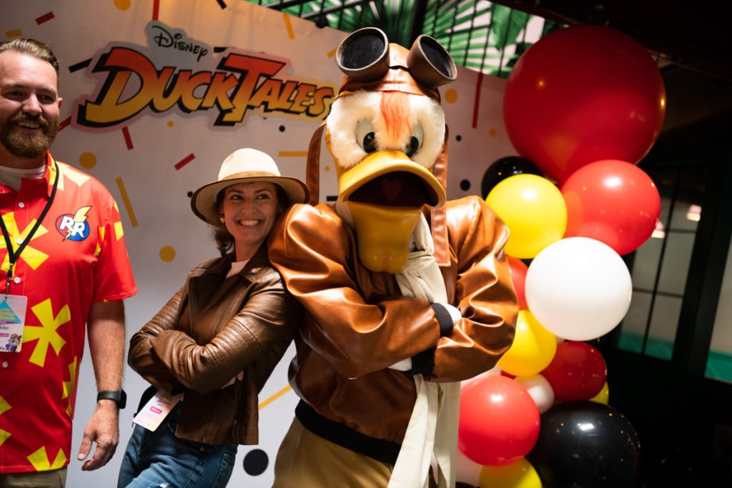 Fan posing with crossed arms back to back with Launchpad McQuack. The fan is dressed as Chip from Rescue Rangers with a white fedora, brown leather jacket, white shirt and blue jeans. Launchpad is tall and muscular with white feathers, orange beak, and tuft of orange hair hanging out of his brown leather pilot’s hat. On top of his hat are his aviator glasses. Launchpad has a brown leather jacket, tan pants, and an off-white scarf. To the left of the fan is a fan smiling with their Dale Rescue Rangers shirt that is a red Hawaiian style shirt with yellow flowers. On the top right corner of the fan’s shirt is the Rescue Rangers logo that is a circle half red and half blue with RR in white block letters and a yellow lightning bolt going through. They are all in front of the photo backdrop that is white with red, yellow and black confetti shapes, there are red, white, yellow, and black balloon towers on either side and a DuckTales logo in the top center of the backdrop.